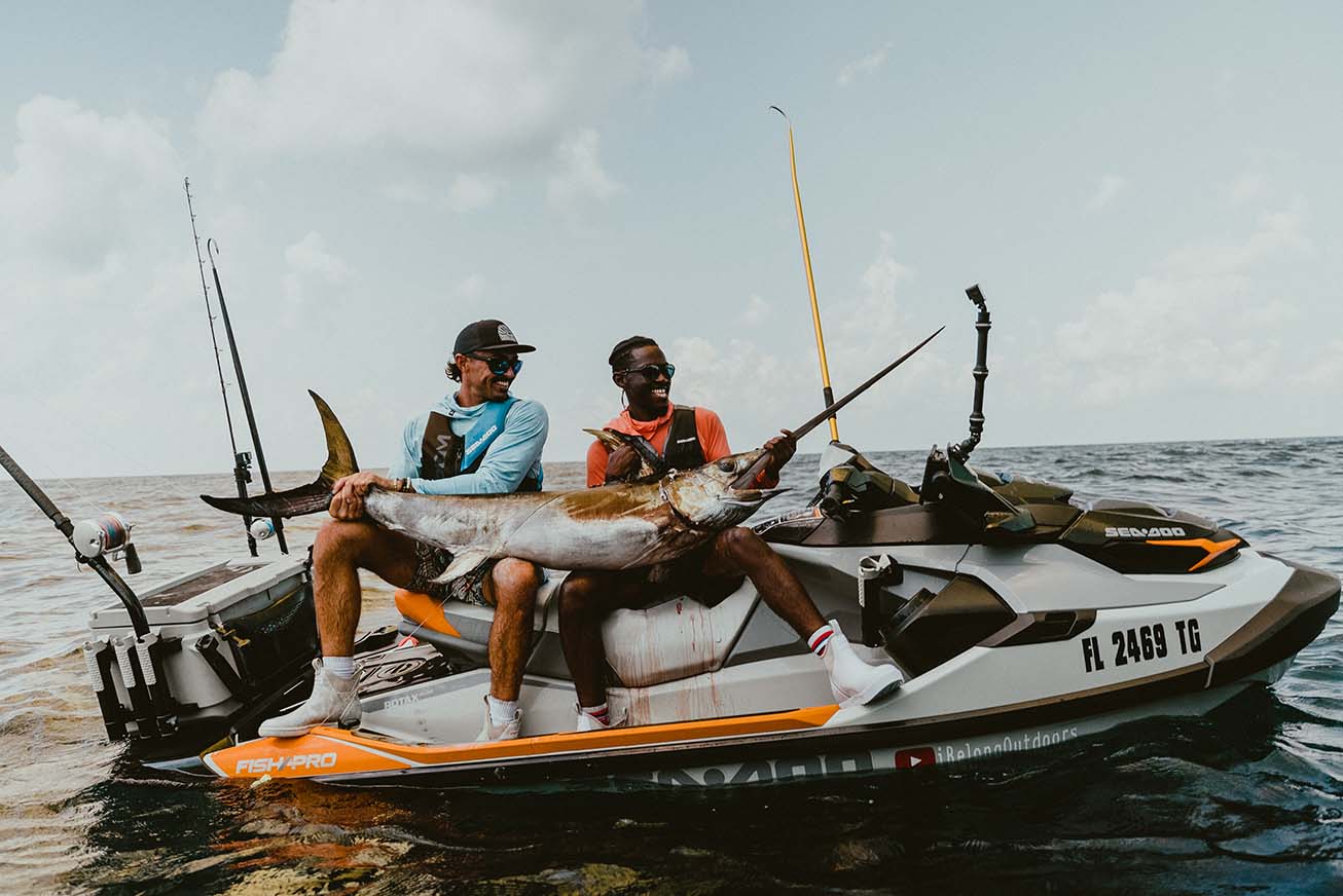 Emmanuel sitting with another man on a jet ski smiling as they hold a large swordfish.