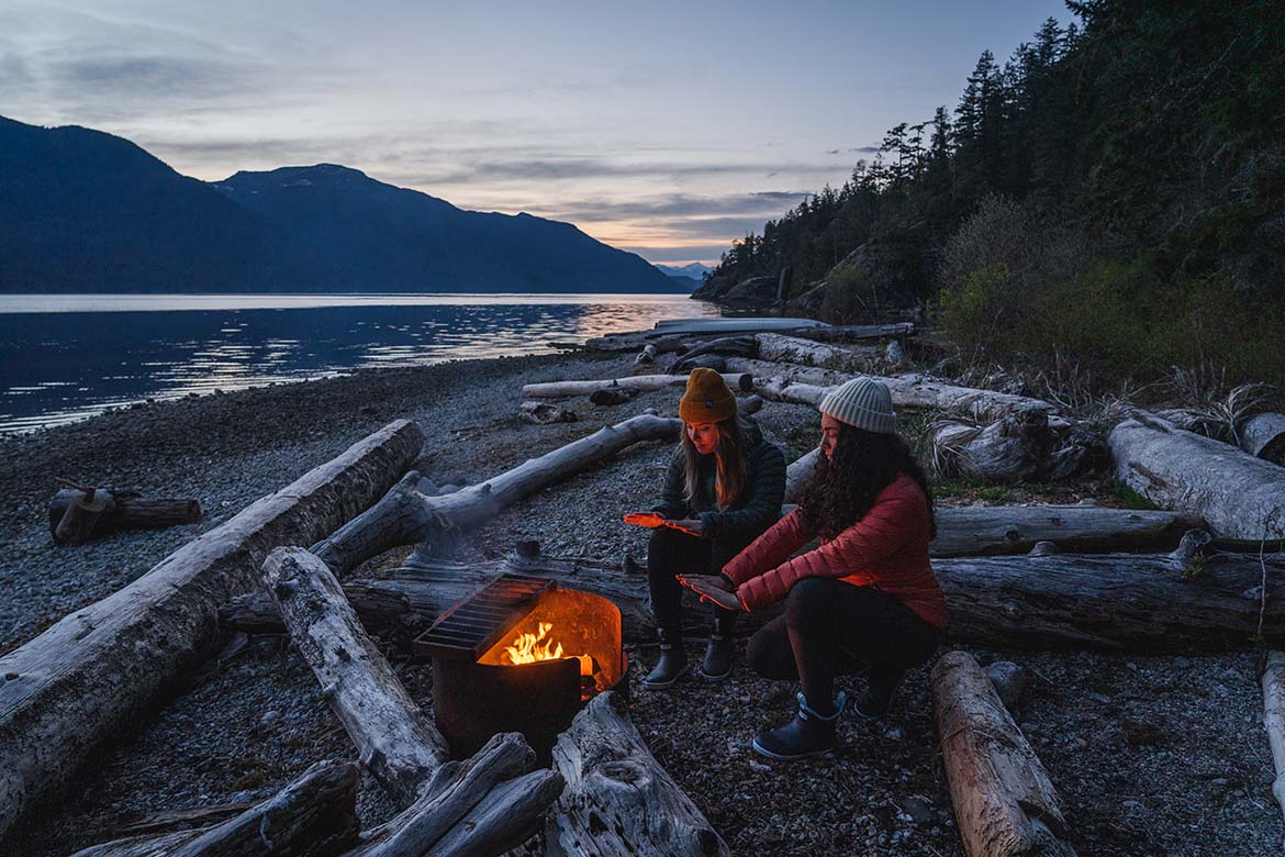 Errin and friend sitting on a log next to a fire water-side